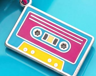 Puede incluir: Un dije de esmalte rectangular con forma de casete, con un borde azul brillante, un panel central magenta y una sección inferior amarilla. El diseño incluye dos carretes circulares, un rectángulo amarillo central y una anilla de color rosa.