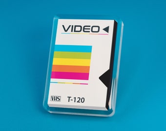 May include: A white VHS video cassette with a black label that says "VIDEO" and a black play button. The label also has a colorful stripe design with blue, green, yellow, orange, and pink stripes. The cassette is labeled "VHS T-120".
