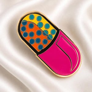 Puede incluir: Un colorido pin de esmalte con forma de pastilla. El pin presenta un borde dorado, un extremo rosa brillante y un interior segmentado con secciones naranjas y amarillas llenas de círculos azules.