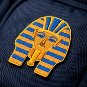 May include: Embroidered patch featuring the golden mask of Tutankhamun, with blue and orange stripes. The patch is sewn onto a navy blue fabric, showcasing the intricate details of the pharaoh's iconic headwear.