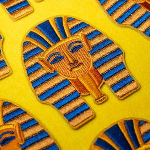 Könnte beinhalten: Gestickte Aufnäher mit der goldenen Maske von Tutanchamun, mit blauen und goldenen Details. Die Aufnäher sind auf einem leuchtend gelben Hintergrund angeordnet und zeigen die aufwendige Stickerei und das Design der altägyptischen Pharaonenmaske.