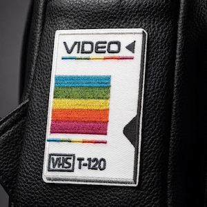 May include: A white embroidered patch featuring the word "VIDEO" above a rainbow design and the text "VHS T-120". The patch is affixed to a black leather surface.