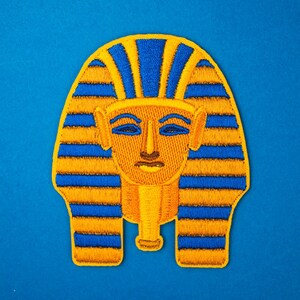May include: Embroidered patch depicting the face of an Egyptian pharaoh, in gold, blue, and brown. The patch shows the pharaoh's headdress and facial features, against a blue backdrop. This patch is suitable for clothing or accessories.