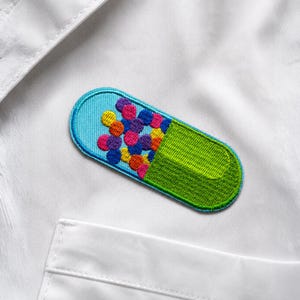Op de afbeelding: Geborduurde patch in de vorm van een capsule, met een lichtblauwe bovenkant en een groene onderkant. De capsule is gevuld met kleurrijke, ronde kralen in tinten geel, roze, paars en oranje. De patch staat op een witte stoffen achtergrond.