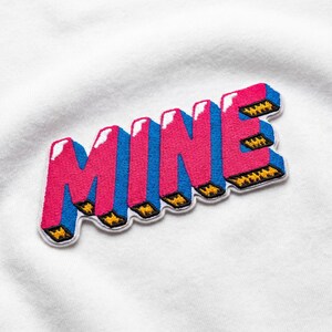 May include: Embroidered patch with the word "MINE" in a 3D bubble letter style. The letters are pink with blue sides and white highlights, outlined in a white border. The patch is on a white fabric background.