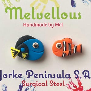 May include: A pair of handmade polymer clay earrings featuring a blue tang fish and an orange clownfish. The earrings are made with surgical steel posts. The text "Melvellous Handmade by Mel" and "Yorke Peninsula S.A. Surgical Steel" is visible in the image.