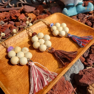 May include: Three wooden bead necklaces with tassels. Each necklace has a different colored tassel: pink and white, brown, and blue and red. The necklaces are displayed on a wooden tray.