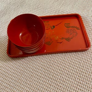 Puede incluir: Un juego de cuatro cuencos lacados rojos apilados sobre una bandeja rectangular a juego. La bandeja presenta un diseño floral dorado. Los cuencos y la bandeja son de color rojo vibrante, lo que sugiere una estética asiática tradicional.