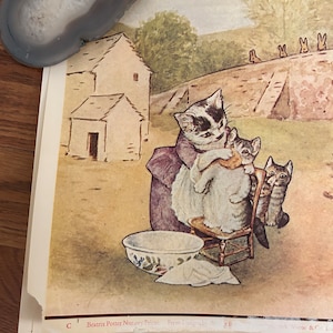 Könnte beinhalten: Ein Vintage-Druck der Beatrix Potter Nursery Frieze mit einer Katze, die eine andere Katze mit einer Schüssel Wasser und einem Handtuch wäscht. Der Druck ist in einem hellbraunen Rahmen mit einem weißen Passepartout. Der Text auf dem Druck lautet "Beatrix Potter Nursery Frieze. From Designs by P. Warne & Co. Ltd."