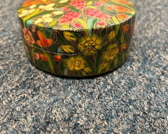 May include: A round, floral-decorated box with a black background. The box is covered in colorful flowers, including pink, yellow, and orange blooms. The box is likely used for storing small items.