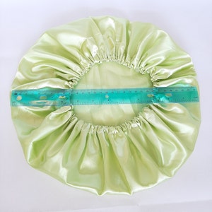 Puede incluir: Un gorro de sat&eacute;n verde claro, de forma circular, con una banda el&aacute;stica. Se coloca una regla sobre el gorro para medir su di&aacute;metro. El gorro est&aacute; hecho de una tela brillante y lisa.