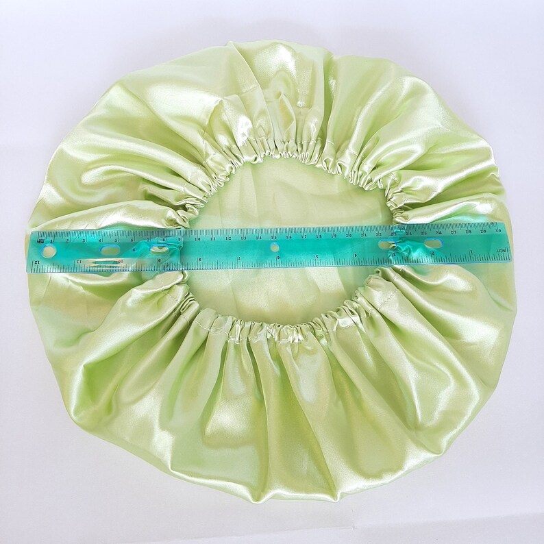 Puede incluir: Un gorro de sat&eacute;n verde claro, de forma circular, con una banda el&aacute;stica. Se coloca una regla sobre el gorro para medir su di&aacute;metro. El gorro est&aacute; hecho de una tela brillante y lisa.