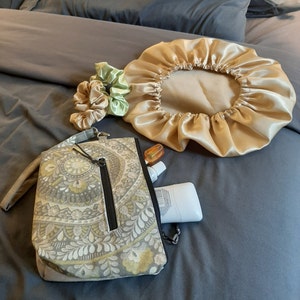 Puede incluir: Una colecci&oacute;n de accesorios de belleza sobre una colcha gris. Incluye un gorro de sat&eacute;n dorado, una bolsa estampada con cremallera negra, coleteros en tonos dorados y verdes y un peque&ntilde;o frasco de protector solar.