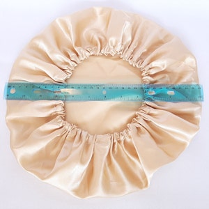 Puede incluir: Un gorro de sat&eacute;n beige con una banda el&aacute;stica, dise&ntilde;ado para proteger el cabello. Una regla azul muestra que el di&aacute;metro del gorro es de aproximadamente 28 cm. El gorro est&aacute; hecho de una tela brillante y suave.