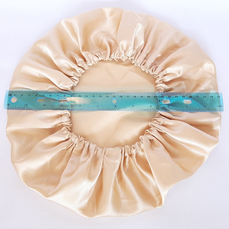 Puede incluir: Un gorro de sat&eacute;n beige con una banda el&aacute;stica, dise&ntilde;ado para proteger el cabello. Una regla azul muestra que el di&aacute;metro del gorro es de aproximadamente 28 cm. El gorro est&aacute; hecho de una tela brillante y suave.