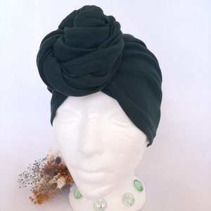 May include: A dark forest green turban hat made of a ribbed fabric. The headwear features a large, prominent knot detail positioned on the front side, creating a gathered and layered appearance that covers the crown and sides of the head.