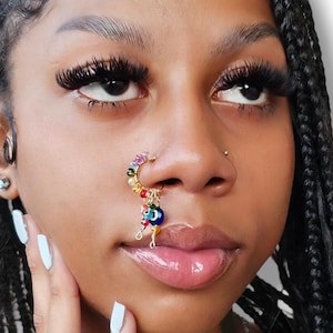 Puede incluir: Primer plano de la cara de una persona con un aro de nariz de cuentas de colores con un amuleto de ojo turco. El aro de nariz está hecho de alambre dorado y tiene una variedad de cuentas, incluyendo azules, verdes, rojas y amarillas. La persona lleva un aro de nariz en la fosa nasal izquierda.