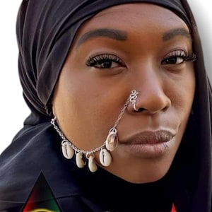 May include: A woman wearing a black headscarf and a red and black jacket with a gold eye symbol. She is wearing a nose chain with seashells hanging from it.