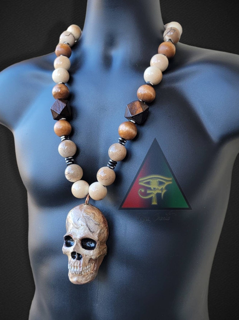 May include: A wooden bead necklace with a skull pendant. The beads are brown, beige, and white. The skull is carved from wood and has a detailed design. The necklace is on a mannequin.