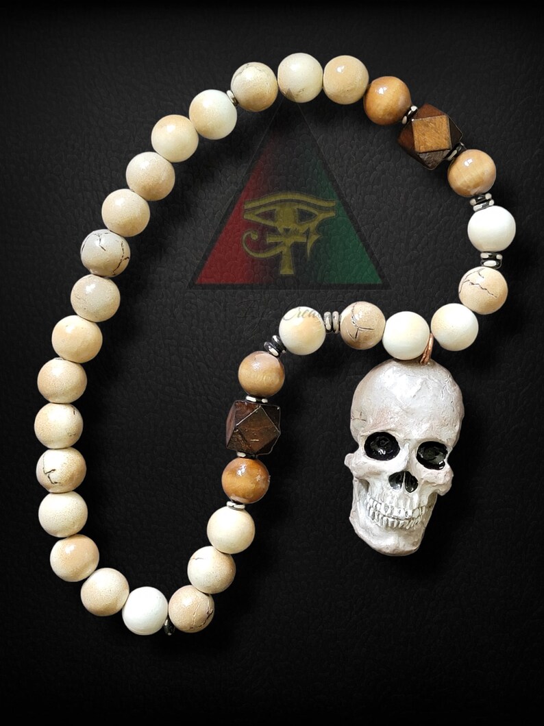 May include: A white and brown beaded necklace with a skull charm. The beads are made of wood and bone. The skull is white with black eyes and a toothy grin. The necklace is hanging on a black background.