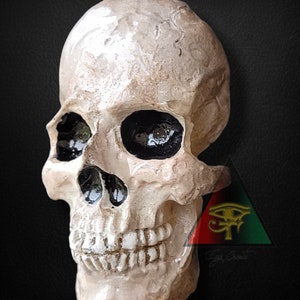 May include: A white ceramic skull pendant with black painted eye sockets and teeth. The skull is hanging from a copper wire loop. The pendant is in front of a red, black, and green triangle with a gold eye of Horus symbol.