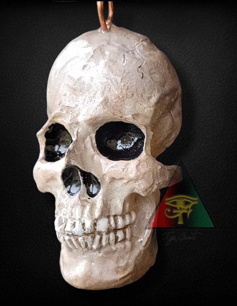 May include: A white ceramic skull pendant with black painted eye sockets and teeth. The skull is hanging from a copper wire loop. The pendant is in front of a red, black, and green triangle with a gold eye of Horus symbol.