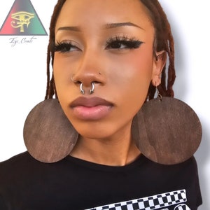 Puede incluir: Una persona lleva grandes pendientes redondos de madera marrón. Los pendientes tienen un diámetro de unos 10 cm. La persona también tiene un piercing en el tabique nasal con un aro plateado.