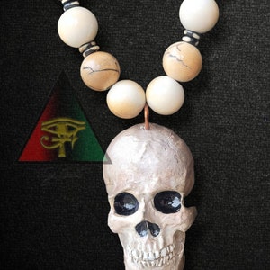 May include: A white skull pendant with black eye sockets hangs from a beaded necklace. The necklace is made of brown and white beads with black accents. The pendant is attached to the necklace with a copper wire.