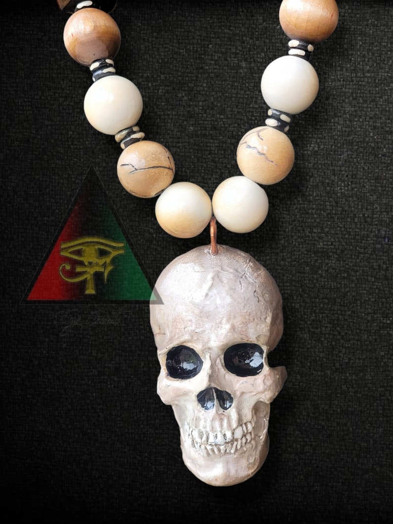 May include: A white skull pendant with black eye sockets hangs from a beaded necklace. The necklace is made of brown and white beads with black accents. The pendant is attached to the necklace with a copper wire.