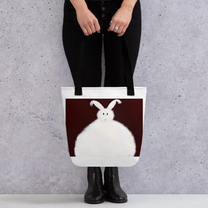 May include: White tote bag with black handles featuring a white fluffy rabbit with long ears on a dark red background.
