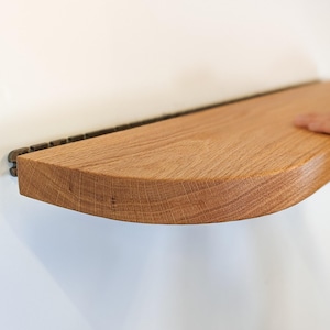 May include: A wooden floating shelf with a curved front edge. The shelf is made of light brown wood and is mounted on a dark metal bracket. The shelf is being held up by a person's hands.