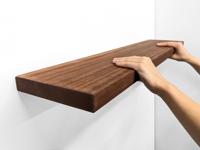 An extra-long, heavy-duty mahogany floating shelf is sucessfully installed and sits securely on a wall. Two hands grip the top and bottom of the front-facing side.