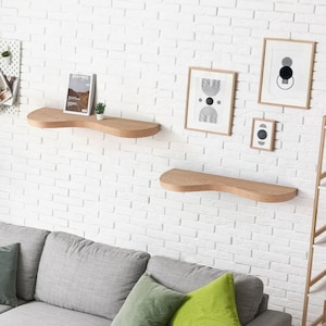 May include: Two wooden shelves with a curved design are mounted on a white brick wall. The shelves are empty except for a small potted plant on the top shelf. There are four framed prints on the wall above the shelves, featuring abstract geometric designs in black and white.