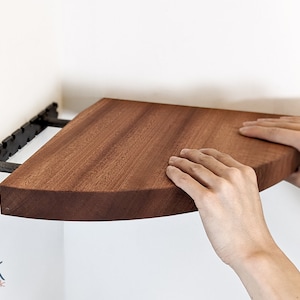 Two hands push a mahogany corner shelf onto the brackets which are wall-mounted.