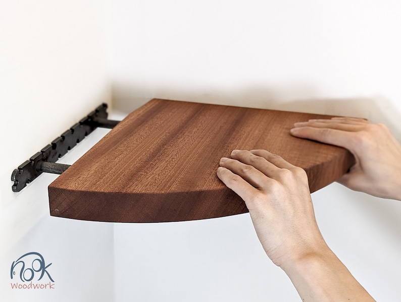 Two hands push a mahogany corner shelf onto the brackets which are wall-mounted.