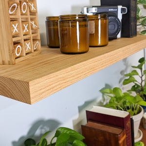 May include: A wooden shelf with three amber glass candle jars, a vintage camera, and a wooden tic-tac-toe game. The shelf is mounted on a white wall with green plants in pots below.