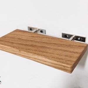 Pu&ograve; includere: Uno scaffale in legno con una finitura marrone chiaro &egrave; montato su una parete bianca usando due staffe metalliche. Lo scaffale &egrave; rettangolare e ha un bordo leggermente arrotondato. Le staffe sono fissate al muro con viti.