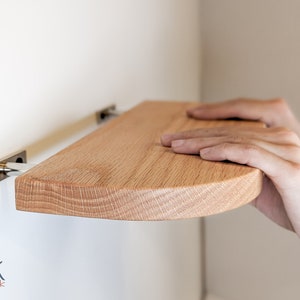 Two hands push a thin oak floating shelf with curved corners onto metal brackets