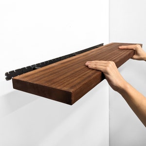 Two hands grip the front of a long wooden mahogany shelf and easily install it on the wall by sliding it onto black brackets.