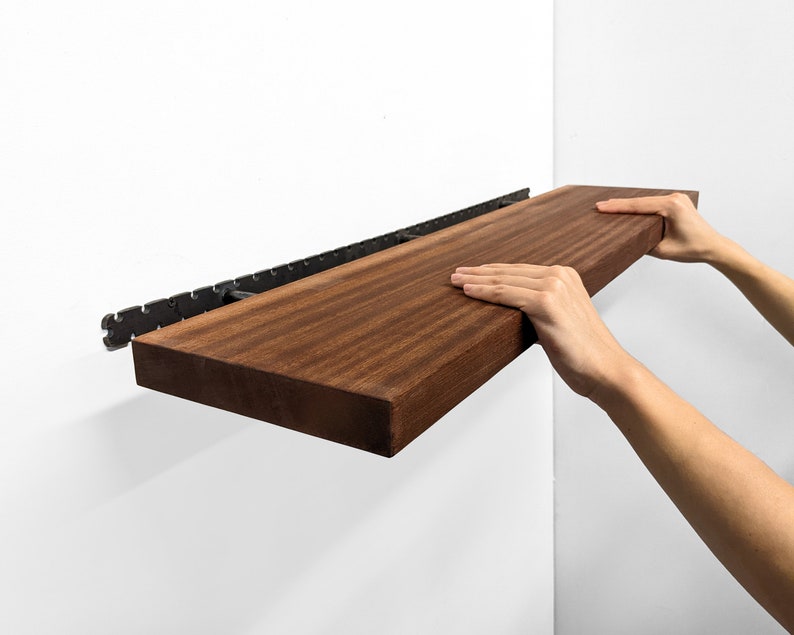 Two hands grip the front of a long wooden mahogany shelf and easily install it on the wall by sliding it onto black brackets.