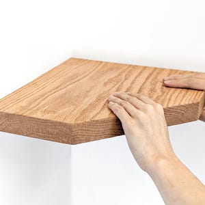 Two hands finish pushing an oak corner floating shelf onto the wall.