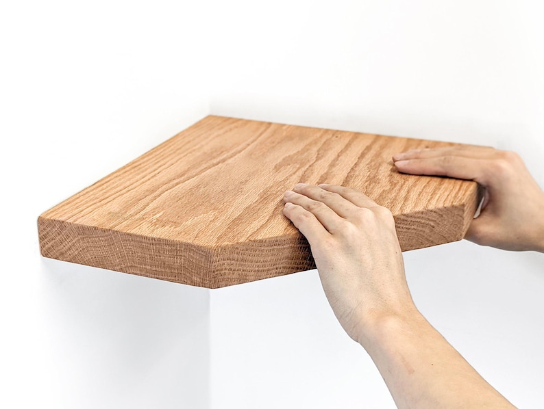Two hands finish pushing an oak corner floating shelf onto the wall.