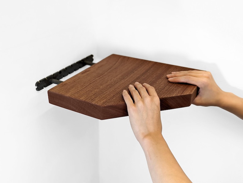Two hands push mahogany corner floating shelf onto wall brackets.