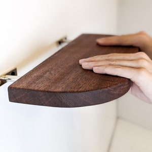 Two hands push a mahogany floating shelf with curved corners onto two metal brackets