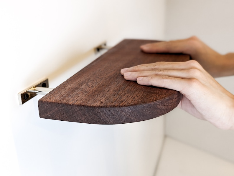 Two hands push a mahogany floating shelf with curved corners onto two metal brackets