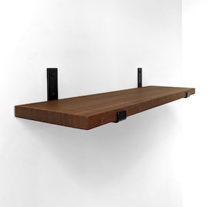 May include: A rectangular wooden shelf with a rich brown wood grain finish, mounted to a wall with two vertical black metal brackets and secured by two smaller black metal clips positioned along the front edge of the shelf surface.