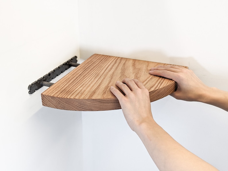 Two hands push a solid wood oak floating corner shelf onto the bracket.
