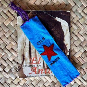 Puede incluir: Un marcapáginas de tela azul con una estrella roja y los textos bordados "EZLN" y "YA BASTA". El marcapáginas tiene una borla morada y está colocado sobre un libro titulado "El Relatos de Anto".
