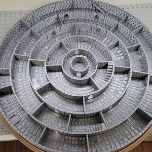 Pode incluir: Um mapa de masmorra circular de vários níveis, cinza e branco, com uma sala circular central. O mapa é feito de papelão e é projetado para jogos de RPG de mesa.