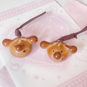 May include: Two brown bear-shaped pendants with dark brown cords. Each pendant has a round nose, small black eyes, and a smiling mouth. The pendants are on a pink and white patterned surface. The text "by CUTEE DOLL" is visible.